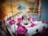 A decorative deity idol adorned with a crown and jewelry is placed in a wooden swing cradle filled with white and pink flowers. Peacock feathers are artistically positioned in the background, enhancing the vibrant and spiritual setup.