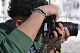 A person with curly hair is holding a DSLR camera up to their eye, preparing to take a photograph. The individual is wearing a green hoodie and several beaded bracelets. The camera strap is visible, and the scene appears to be outdoors with sunlight creating shadows.