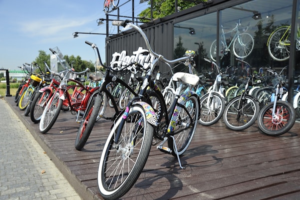 A collection of colorful bicycles is displayed on a wooden platform outside a glass-fronted building. The bicycles are arranged in a row and include various designs and colors, from black and red to multicolored patterns. The background shows reflections of additional bicycles inside the building, along with trees and a clear sky.