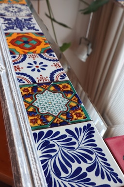A close-up view of a decorative strip featuring a series of colorful ceramic tiles. Each tile displays intricate geometric and floral patterns in vivid colors such as blue, orange, and green. The tiles are arranged in a linear sequence, bordered by a silver frame. In the background, there are blurred elements of indoor decor, including a soft-colored curtain and a plant with green leaves.