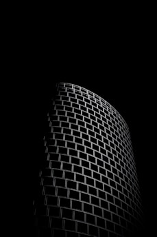 A dark, dramatic image highlighting the curved facade of a modern building. The structure features a repetitive geometric pattern of rectangular windows, sharply contrasted against a black background.