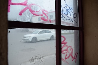 A view through a graffiti-covered window showing a white car driving on a street outside. The window features pink and black graffiti tags while the background includes a blurry building.