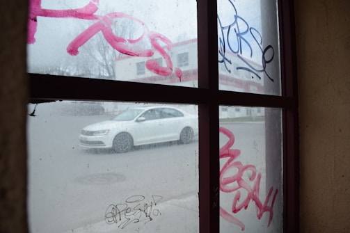 A view through a graffiti-covered window showing a white car driving on a street outside. The window features pink and black graffiti tags while the background includes a blurry building.
