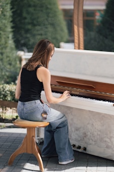 A young woman sits on a wooden stool while playing an upright piano outdoors. She wears a black sleeveless top and casual jeans. The setting appears to be a garden or park with greenery in the background. A keychain hangs from her jeans pocket.