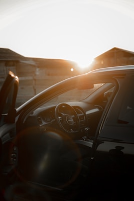 A black car with an open door, showcasing a detailed view of the interior. The focus is on the steering wheel, which features a logo. The scene is set during sunset, with the sunlight creating a warm glow and lens flare effect. Wooden structures are visible in the background.