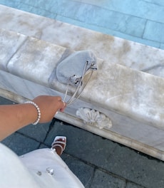 A person is dressed elegantly, holding a small checkered bag while standing near a marble surface with intricate floral carvings. The person is wearing a bracelet and white sandals, suggesting a stylish and sophisticated setting.
