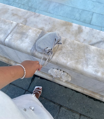 A person is dressed elegantly, holding a small checkered bag while standing near a marble surface with intricate floral carvings. The person is wearing a bracelet and white sandals, suggesting a stylish and sophisticated setting.