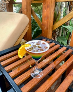 A colorful cocktail in a martini glass is placed on a wooden table. The drink features a gradient of vibrant hues topped with a layer of ice cubes and is garnished with a slice of orange. The background consists of wooden beams and lush greenery, giving an outdoor and natural ambiance.