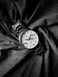 A silver wristwatch with a metal strap lies elegantly on a backdrop of dark, textured fabric. The watch face is classic, featuring Roman numerals and two smaller dials for additional functionalities. The design is sophisticated and minimalist, with a small red logo adding a subtle accent to the otherwise monochrome palette.