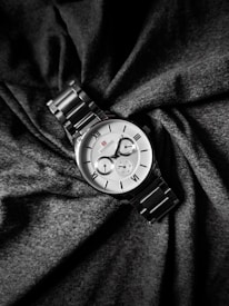 A silver wristwatch with a metal strap lies elegantly on a backdrop of dark, textured fabric. The watch face is classic, featuring Roman numerals and two smaller dials for additional functionalities. The design is sophisticated and minimalist, with a small red logo adding a subtle accent to the otherwise monochrome palette.