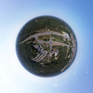 A spherical aerial view of a landscape featuring roads, greenery, and buildings, creating a planet-like effect. The central area is densely covered with forest, surrounded by winding roads and some residential and commercial buildings.