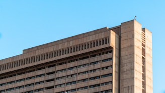 A modern, high-rise concrete building with multiple windows under a clear blue sky. The structure is characterized by a minimalist design with clean lines and a uniform facade.