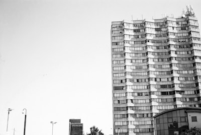 A tall, multi-story building with a unique wavy facade stands against a clear sky. The building has many windows and an array of antennas on the roof. Nearby, there are street lights and a smaller structure labeled 'Dreamland.' Trees and other urban elements are visible at the bottom of the frame.