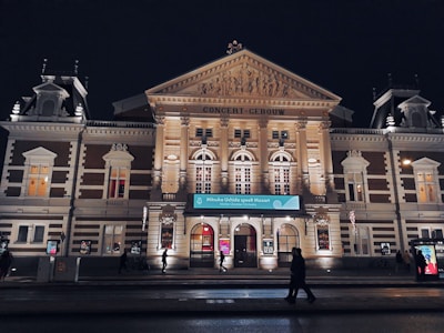 A grand, historic concert hall is illuminated at night, showcasing ornate architectural details with columns and decorative sculptures. People are walking by, and a banner advertises a performance.