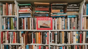 A densely packed bookshelf full of various books, with titles spanning multiple genres and topics. The centerpiece is a prominently displayed red book titled 'Titanic,' featuring an image of the ship. The books are of varying sizes and colors, showcasing a wide array of literary interests.