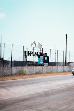 A roadside scene featuring a fenced area with steel structures and two tilted advertising billboards. The billboards display a QR code and some text. The sky is clear and the road runs alongside the fence, with a small portion of a vehicle visible on the right.