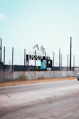 A roadside scene featuring a fenced area with steel structures and two tilted advertising billboards. The billboards display a QR code and some text. The sky is clear and the road runs alongside the fence, with a small portion of a vehicle visible on the right.
