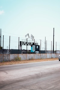 A roadside scene featuring a fenced area with steel structures and two tilted advertising billboards. The billboards display a QR code and some text. The sky is clear and the road runs alongside the fence, with a small portion of a vehicle visible on the right.