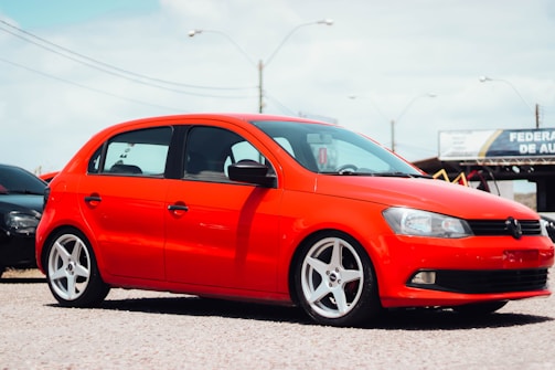 A bright red compact car is parked on a gravel surface. It has sleek, silver alloy wheels and tinted windows. In the background, another car and a sign for an auto business are visible.