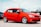 A bright red compact car is parked on a gravel surface. It has sleek, silver alloy wheels and tinted windows. In the background, another car and a sign for an auto business are visible.