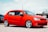 A bright red compact car is parked on a gravel surface. It has sleek, silver alloy wheels and tinted windows. In the background, another car and a sign for an auto business are visible.