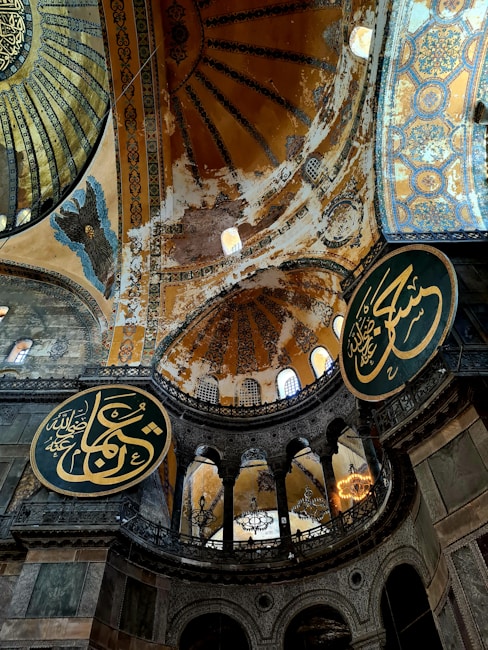 A grand architectural interior with ornate, intricate designs and calligraphy. The structure features large circular plaques with Arabic script, an expansive dome with intricate geometric patterns, and muted colors accented by golds and blues. The aged walls exhibit a rustic look, enhancing the historic ambiance. Wrought iron chandeliers hang from the ornate ceiling.