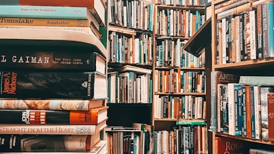 A densely packed library filled with books on wooden shelves. The image prominently features stacks of books in the foreground with various titles, while more shelves filled with books recede into the background. The scene is rich with literature and conveys an atmosphere of knowledge.