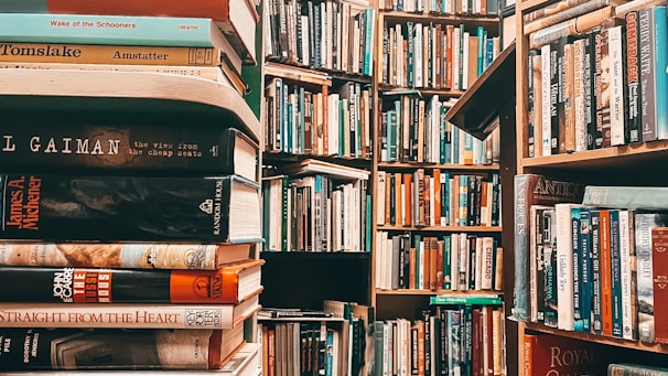 A densely packed library filled with books on wooden shelves. The image prominently features stacks of books in the foreground with various titles, while more shelves filled with books recede into the background. The scene is rich with literature and conveys an atmosphere of knowledge.