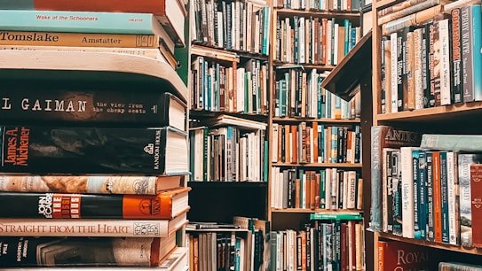 A densely packed library filled with books on wooden shelves. The image prominently features stacks of books in the foreground with various titles, while more shelves filled with books recede into the background. The scene is rich with literature and conveys an atmosphere of knowledge.