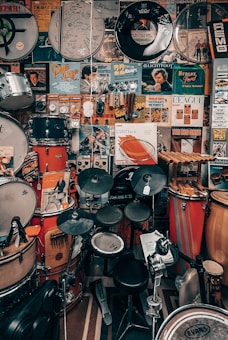 A room filled with an eclectic collection of musical memorabilia, including various types of drums, concert posters, and signed drumheads. The walls are adorned with vibrant posters featuring musicians and bands. Several drum kits and percussion instruments are neatly arranged, occupying most of the space.