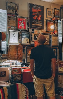 A person stands in a room filled with framed posters and pictures on the walls. The room has a vintage or retro decor, with various prints and posters related to music and entertainment. The person appears to be examining the wall of artwork, which includes imagery of musical instruments and performers. The ambiance is warm and somewhat nostalgic, created by the lighting and color tones.