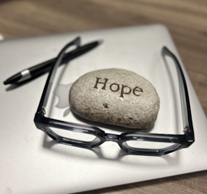 A smooth stone inscribed with the word 'Hope' sits on top of a closed laptop. Next to it are a pair of black-framed glasses and a black pen, all resting on a wooden surface.