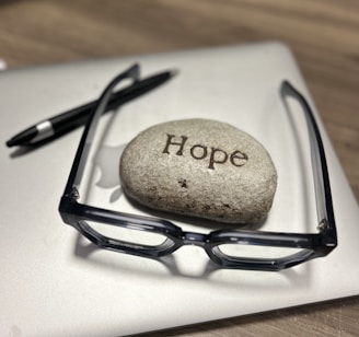 A smooth stone inscribed with the word 'Hope' sits on top of a closed laptop. Next to it are a pair of black-framed glasses and a black pen, all resting on a wooden surface.