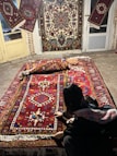 A person is kneeling next to a collection of intricately designed Persian rugs with vibrant patterns and colors. The rugs are displayed on a stone floor and hanging on doors, each showcasing a variety of geometric and floral motifs.