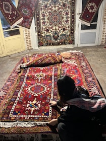 A person is kneeling next to a collection of intricately designed Persian rugs with vibrant patterns and colors. The rugs are displayed on a stone floor and hanging on doors, each showcasing a variety of geometric and floral motifs.