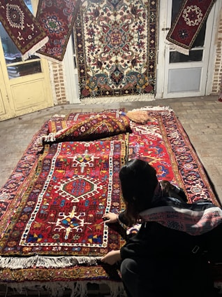A person is kneeling next to a collection of intricately designed Persian rugs with vibrant patterns and colors. The rugs are displayed on a stone floor and hanging on doors, each showcasing a variety of geometric and floral motifs.