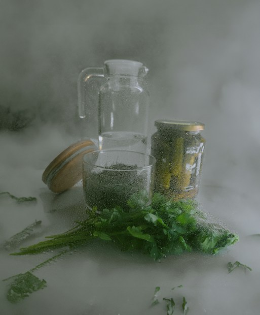 A misty scene featuring a transparent glass pitcher, a jar of pickles, and a bunch of fresh green herbs placed on a surface. The surfaces are covered in condensation, adding a foggy appearance to the overall composition.
