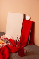 A blank canvas is propped against a wall, surrounded by red fabric. Several paintbrushes are placed in a glass jar, and a book with colorful illustrations is partially covered by the fabric. There's also a red transparent mug in the foreground.