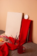 A blank canvas is propped against a wall, surrounded by red fabric. Several paintbrushes are placed in a glass jar, and a book with colorful illustrations is partially covered by the fabric. There's also a red transparent mug in the foreground.