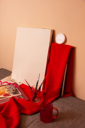 A blank canvas is propped against a wall, surrounded by red fabric. Several paintbrushes are placed in a glass jar, and a book with colorful illustrations is partially covered by the fabric. There's also a red transparent mug in the foreground.
