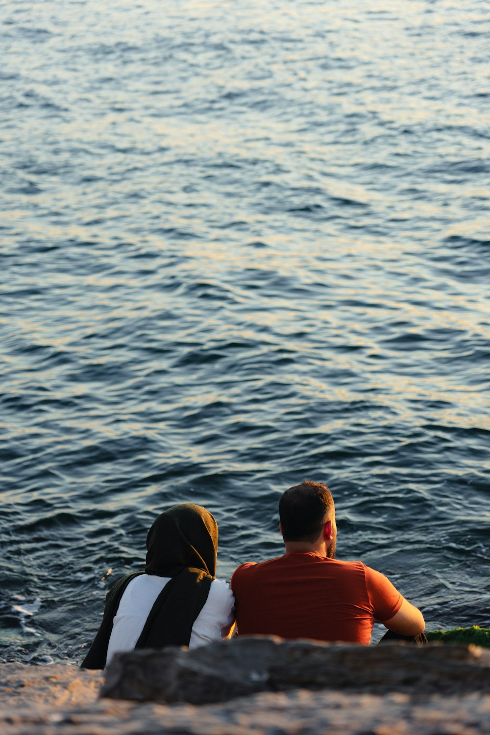 um casal de pessoas sentadas em cima de uma praia ao lado de um corpo de água