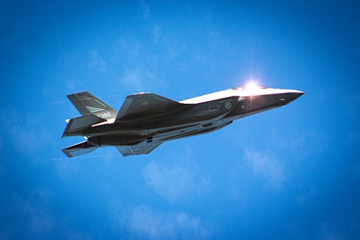 A military jet is soaring through a clear blue sky, highlighted by sunlight reflecting off its surface. The aircraft features a sleek, angular design, with visible markings and insignia on its body.