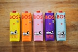 Five colorful cartons of BOS iced tea are arranged on a wooden surface. Each carton has a unique color corresponding to its flavor: yellow for lemon, orange for peach, pink for watermelon and mint, purple for berry, and blue for lime and ginger. The cartons feature the BOS logo in bold red letters and an illustration of a lion lounging.