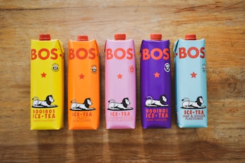 Five colorful cartons of BOS iced tea are arranged on a wooden surface. Each carton has a unique color corresponding to its flavor: yellow for lemon, orange for peach, pink for watermelon and mint, purple for berry, and blue for lime and ginger. The cartons feature the BOS logo in bold red letters and an illustration of a lion lounging.