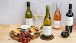 A selection of wine bottles including white, rosé, and red wine, arranged on a wooden table. There is a round wooden serving board with red grapes, crackers, a tub of cheese spread, and a knife. Two wine glasses are also present, one filled with white wine and the other with red wine. Green olives are placed in a small bowl. The background features a light, textured wall.