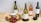 A selection of wine bottles including white, rosé, and red wine, arranged on a wooden table. There is a round wooden serving board with red grapes, crackers, a tub of cheese spread, and a knife. Two wine glasses are also present, one filled with white wine and the other with red wine. Green olives are placed in a small bowl. The background features a light, textured wall.