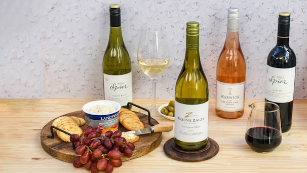 A selection of wine bottles including white, rosé, and red wine, arranged on a wooden table. There is a round wooden serving board with red grapes, crackers, a tub of cheese spread, and a knife. Two wine glasses are also present, one filled with white wine and the other with red wine. Green olives are placed in a small bowl. The background features a light, textured wall.