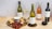 A selection of wine bottles including white, rosé, and red wine, arranged on a wooden table. There is a round wooden serving board with red grapes, crackers, a tub of cheese spread, and a knife. Two wine glasses are also present, one filled with white wine and the other with red wine. Green olives are placed in a small bowl. The background features a light, textured wall.