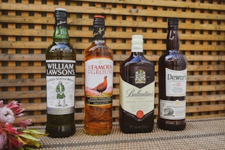 Four bottles of blended Scotch whisky are arranged on a textured surface with a wooden lattice background. The bottles include William Lawson's, The Famous Grouse, Ballantine's, and Dewar's. To the left, there is a small arrangement of flowers.