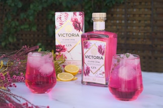 A bottle of Victoria Classic Pink Gin is placed on a marble surface, surrounded by floral decorations and slices of lemon. Two glasses filled with pink gin and ice cubes are garnished with herbs. The background features a wooden lattice and green foliage, giving a fresh and inviting atmosphere.